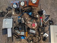 Photo showing a mixed lot of electronics and accessories including an Apple TV, spotlights, remotes, chargers, cables and electronic parts on carpeted floor.