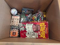 Photo showing an open box containing various toys including a baseball, a vintage electronic diffusion detector kit in original box, mini lamp boxes, and other items.