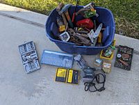 Blue plastic tub filled with various hand tools including hammers, wrenches, pliers, hand saws. Several drill bit sets laid out nearby including Dewalt and other brands. Black+Decker orbital sander with power cord visible.