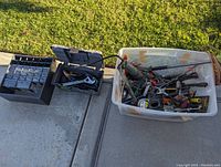 Wide view showing large plastic tub of assorted hand tools, black parts organizer and two small toolboxes on concrete surface.