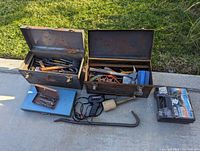 Two metal toolboxes open displaying assorted hand tools, including hammers, pliers, screwdrivers, and more, set on concrete next to grass
