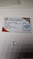 Photo showing the Chanthaburi Gemological Laboratory certification card with details of the aquamarine gemstone and the loose pear drop shaped gemstone on white background.