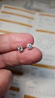 Photo showing both moissanite earrings held by fingers, focus on stones and setting with certification papers visible in background.