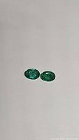 Photo showing two oval shaped Zambian emerald gemstones placed on a plain surface highlighting their green color and luster.