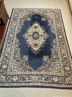 Full view of rectangular area rug laid on carpet, showing blue field with central beige floral medallion and decorative borders