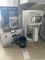 Photo of coffee maker on counter with both boxed coffee grinders and black CKCO mug nearby