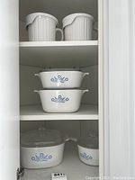 Three white CorningWare casserole dishes with blue cornflower pattern and glass lids, plus one white saucepan with matching lid, and two soup cups with lids on shelves.