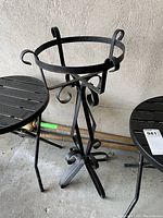 Black metal plant stand with decorative scrollwork shown beside two black metal folding tables.