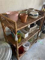 Rattan shelf unit with various planter pots and other items on shelves (contents not included in lot). Shows woven rattan surface and tubular rattan frame construction.