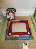 Photo showing the small MAD tin lunch box and boxed Etch A Sketch side by side on carpet