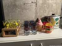 Photo showing six lemon-patterned glasses in a wooden carrier, Pepsi-Cola glasses, various plastic cups, and a large floral pot.