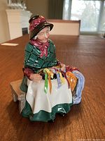 Front view of the Royal Doulton figurine showing seated elderly woman sewing ribbons on white cloth while sitting on a bench.