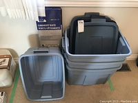 Photo showing three large gray Rubbermaid storage containers stacked together with matching lids, one smaller lid on top, and a boxed garment rack behind them.