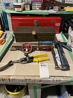 Red metal toolbox in background, brown metal removable shelf/toolbox with lid open showing hand tools inside on table in front. Included vintage hand drill with red handle, caulking gun, bolt cutters with yellow handles.