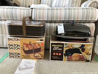 Salton hot bread basket with original box and instructions alongside electric hot server with original box, placed on carpet with striped couch background.