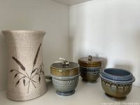 Four items on a white shelf: one ceramic vase with brown leaf designs, three Wade clover jars, two with lids and one without.