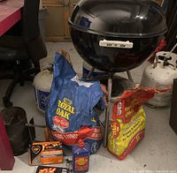 Photo showing Weber kettle BBQ grill with two bags of Royal Oak charcoal, charcoal chimney starter, Zip fire starters box, blue fire starter fluid bottle, and three white propane cylinders grouped around grill