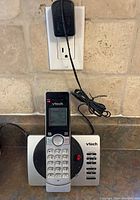 Single cordless handset placed on base, showing numeric keypad, small LCD screen and base power adapter connected to outlet