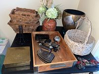 Overview of various decorative items including three stacked square wicker baskets, ceramic vase with faux flowers, glazed pottery pot on glass base, rectangular wooden tray, metal hinged box, metal apple figurine, cast iron iron, miniature limousine model, and motorcycle decor.