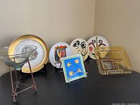 Overview of lot including six decorative items: ceramic plates with abstract face designs, blue tile, glass bowl on metal stand, and 22K gold plated square plate