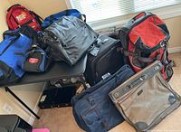 Assorted luggage bags piled on and around a table near a window, showing various types and colors including backpacks, gym bags, and suit bags.