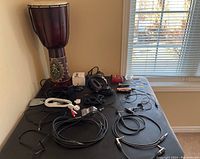 Photo of music accessories on a black table including the wooden djembe drum, black headphones, guitar picks, instrument cables, power adapters and pedals.