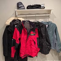 Seven outdoor clothing pieces hanging on hooks including a black Beaver Canoe parka, a red and black Gore-Tex jacket, and other jackets and pants.