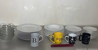 Stack of white ceramic plates and bowls with ribbed design, several assorted coffee mugs, and a set of 4 clear glass bowls