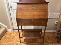 Front view of vintage wood secretary desk with closed drop-front panel, brass hardware, tapered legs, and lower shelf.