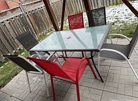 Photo of rectangular patio table with textured tempered glass top and six sling chairs around it in black, red, and grey colors, on outdoor patio.