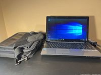 Photo showing Toshiba laptop powered on displaying Windows 10 home screen, with Targus soft case beside it on a black table.