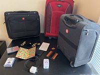 Three roller bags are displayed on a table including a black Swiss Gear bag, a red Skyway bag, and a gray Air Canada carry-on. Travel universal adapter, deck of cards, travel Scrabble game, and travel backgammon game pieces and boards are also laid out.