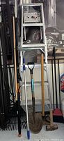 Photo showing the metal stepladder with several tools leaning against it including the Fiskars tree pruner, shovel, rake, and broom.