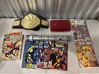 Wide view of the lot showing comic books spread out, championship wrestling belt, Star Wars lightsaber replica, and a red plastic case on a white table.