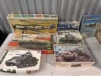 Seven military model kits displayed on a table, including multiple airplane and tank kits plus a battleship kit in their original boxes.