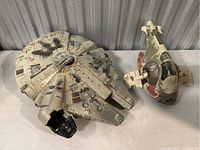 Two Star Wars ships side by side on a grey surface with a corrugated metal background, showing the Millennium Falcon and Slave 1 models.