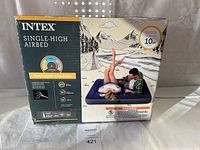 Front of box showing air bed brand (Intex), type (Single-High Airbed), size (Queen), height (10 inches), and a photo of two people sitting on the inflated air bed outdoors.