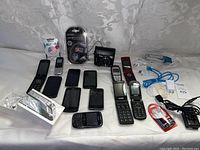 Photo showing multiple used phones including Apple iPhones, Blackberry phone, Motorola flip phones, cables, chargers, and Samsung camera with case.