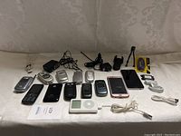 Photo showing an assortment of old mobile phones laid out on a flat surface including Motorola, Samsung, LG, and iPhones, plus an Apple iPod Classic, several charging cables, wired earphones, and phone accessories
