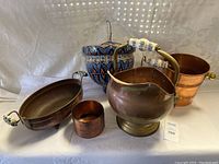Photo showing copper kettle with porcelain handles, small round copper container, oval copper dish with porcelain handles, small copper pot and ceramic hanging planter in background.