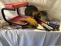 Image shows red Griot's Garage creeper seat with black metal frame, yellow Tillman 495L gloves, large bolt cutters, wood hand saw, and various hand tools arranged on a white cloth.