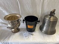 Three ice buckets on white fabric background: ornate silver grapevine design bucket, black metal champagne cooler, and pewter bell-shaped bucket.