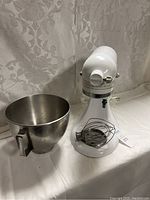 White KitchenAid stand mixer with stainless steel bowl and wire whisk attachment on a white surface