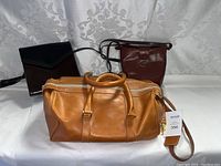 The full lot showing three leather purses on a white background: tan brown duffel bag front and center, dark brown crossbody bag to the right, black clutch to the left.