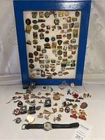 Full display of framed pins in blue frame with multiple loose pins, cufflinks, and wristwatch laid out below on a white surface.