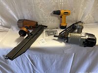 All three tools displayed together from a front angle on white cloth background, including two Bostitch nail guns and one yellow DeWalt drill with manuals