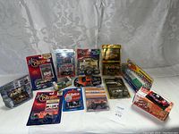 Photo of collection of approximately 14 assorted die-cast racing cars and truck models, mostly in original packaging, laid out on white fabric background.