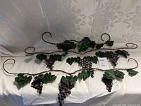Three stained glass grapevine wall hangings laid side by side on a white textured fabric background, showing purple and green grape clusters and green leaves with metal vine structure and hooks.