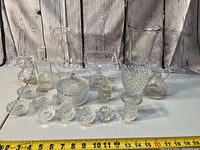 Photo showing full lot of clear glass and crystal glassware items including pitchers, vases, dishes and small holders on white surface with measuring tape for size context.