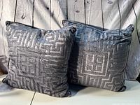 Two large grey velvet pillows with geometric raised pattern, placed in front of a white wood textured background.